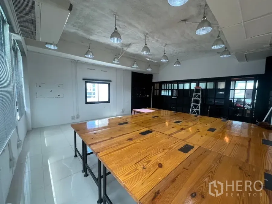 Office Space for rent in Khlong Toei, Bangkok - Open-plan office with large wooden worktables, pendant lights, AC units and a black-framed glass partition.