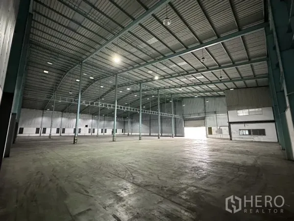 Warehouse for rent in Mueang Samut Prakan, Samut Prakan - Open warehouse floor with high clearance, skylights, roll-up door and a small built-in office section.