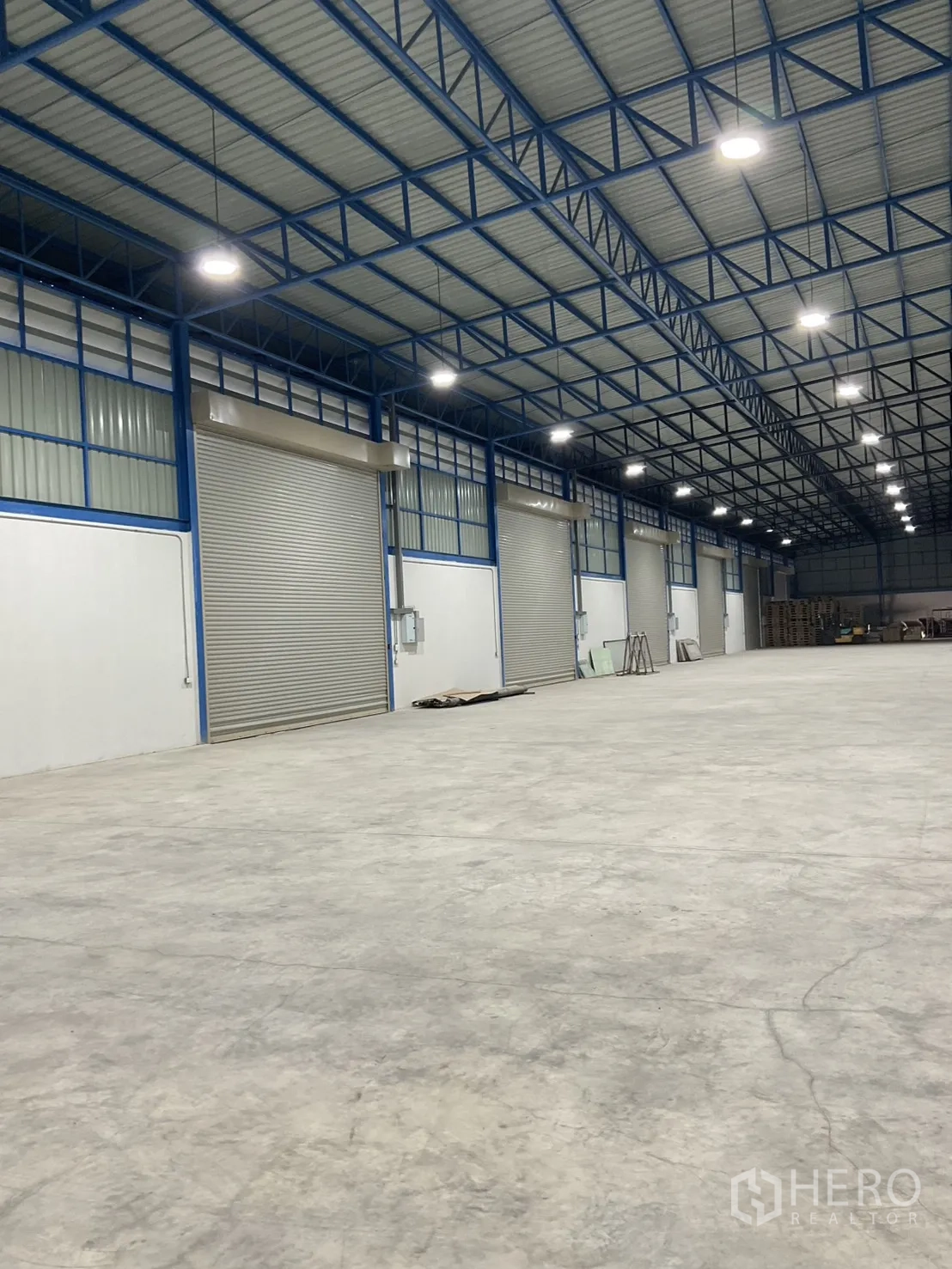 Warehouse for rent in Bang Bua Thong, Nonthaburi - High-clear warehouse interior with steel truss roof, bright LED lights, and several roller shutters.