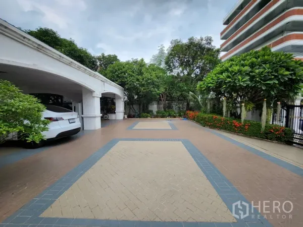 Detached House for rent in Prawet, Bangkok - Spacious tiled forecourt with landscaped garden borders and multiple‑car covered parking beside the house.