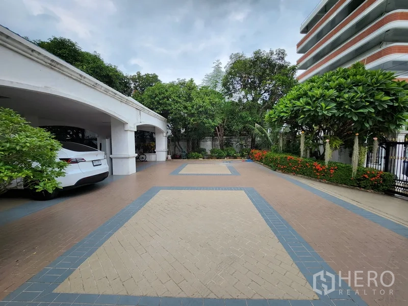 Detached House for rent in Prawet, Bangkok - Spacious tiled forecourt with landscaped garden borders and multiple‑car covered parking beside the house.