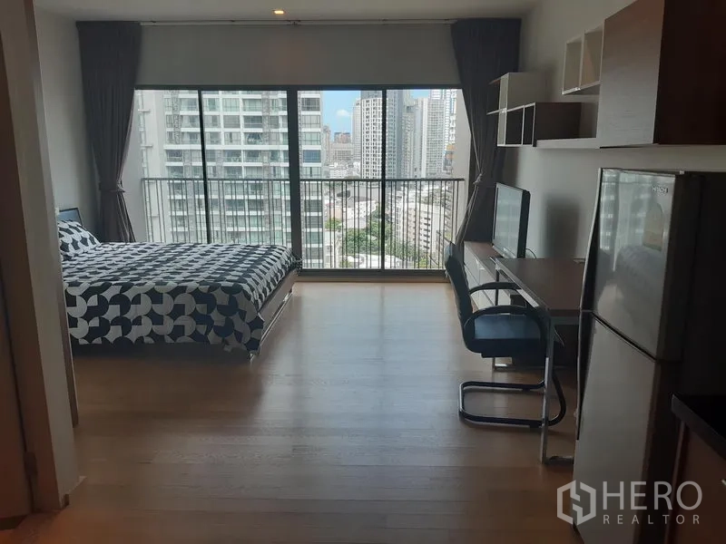 Condominium for rent in Bangkok - Bright studio condo with floor-to-ceiling windows, bed, desk and a city skyline view from the balcony.