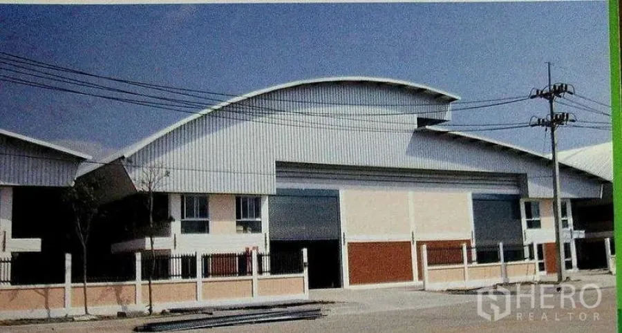 Factory for rent in Ban Phaeo, Samut Sakhon - Factory exterior with curved metal roof, large roller shutter door, and fenced frontage in an industrial street.