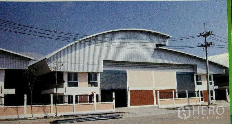 Factory for rent in Ban Phaeo, Samut Sakhon - Factory exterior with curved metal roof, large roller shutter door, and fenced frontage in an industrial street.