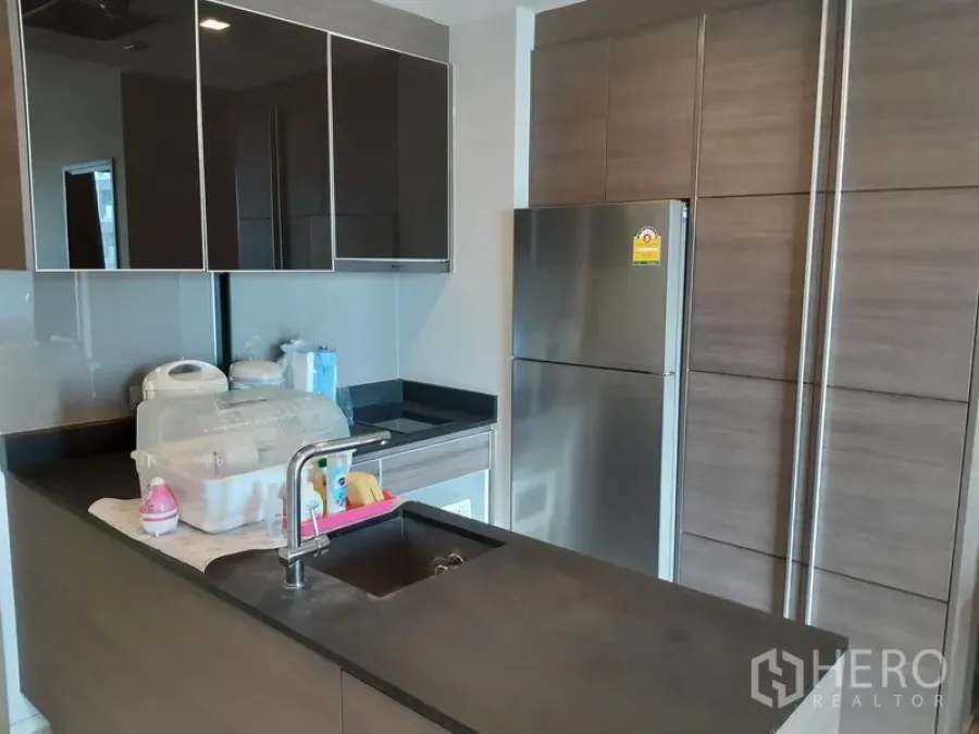 Condominium for rent in Khlong Toei, Bangkok - Modern kitchenette with dark countertops, glossy cabinets and a stainless-steel refrigerator.