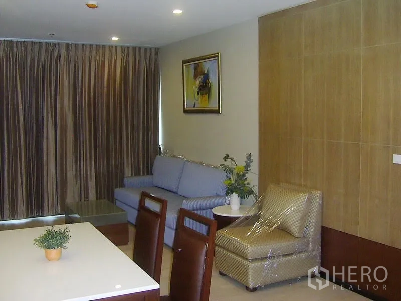 Condominium for rent in Khlong Toei, Bangkok - Open-plan Khlong Toei condo living room with blue sofa, armchair, wood panel wall and floor-to-ceiling curtains.