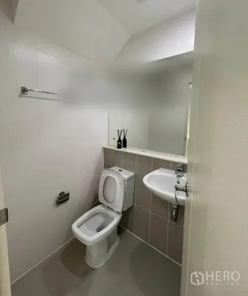 Townhouse for sale in Suan Luang, Bangkok - Clean guest bathroom with white tiles, toilet, wall mirror and corner sink.
