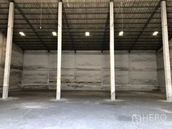 Warehouse for rent in Lat Krabang, Bangkok - Spacious warehouse interior with high ceiling, steel trusses, concrete floor and central support columns illuminated by overhead lights.