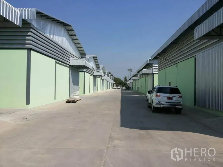 Warehouse for rent in Lam Luk Ka, Pathum Thani - Row of modern mint‑green warehouse units with metal roofs and a wide concrete access road for trucks.
