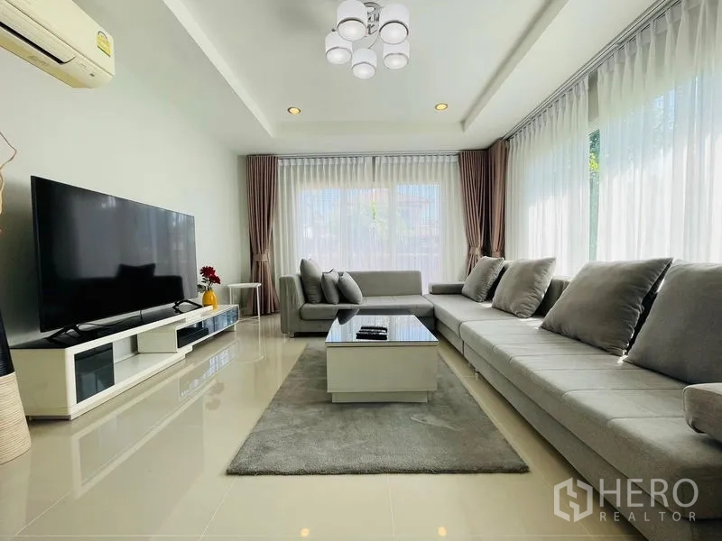 Detached House for sale in Lam Luk Ka, Pathum Thani - Spacious family lounge with long sectional sofa, large TV and full-height windows.