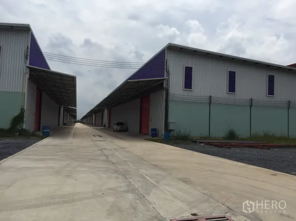 货栈 出租在Pathum Thani - 两侧仓库对向而立，带雨棚与红色装卸门，中间混凝土道路。