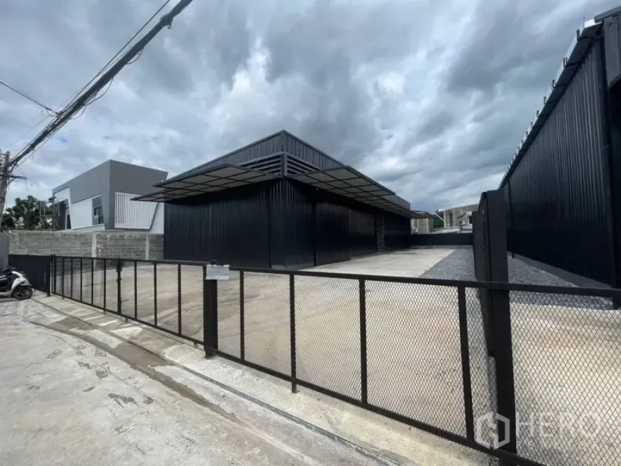 Warehouse for rent in Lat Phrao, Bangkok - Gated entrance and large concrete yard in front of a steel-clad warehouse with a wide loading canopy.