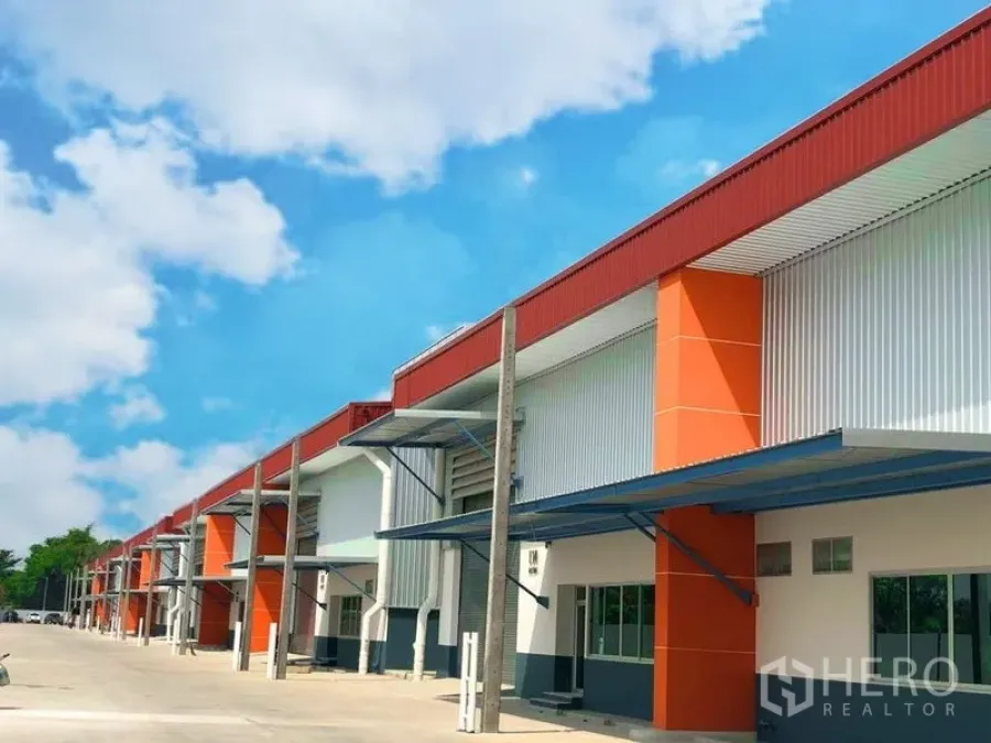 Factory for rent in Bang Phli, Samut Prakan - Row of modern factory-warehouse units with orange accents, covered loading bays and a wide driveway under a blue sky.