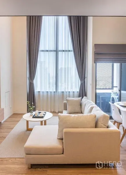 Condominium for rent in Khlong Toei, Bangkok - L‑shaped sofa facing floor‑to‑ceiling windows with sheer curtains and city light.