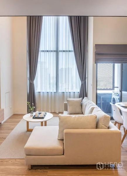 Condominium for rent in Khlong Toei, Bangkok - L‑shaped sofa facing floor‑to‑ceiling windows with sheer curtains and city light.