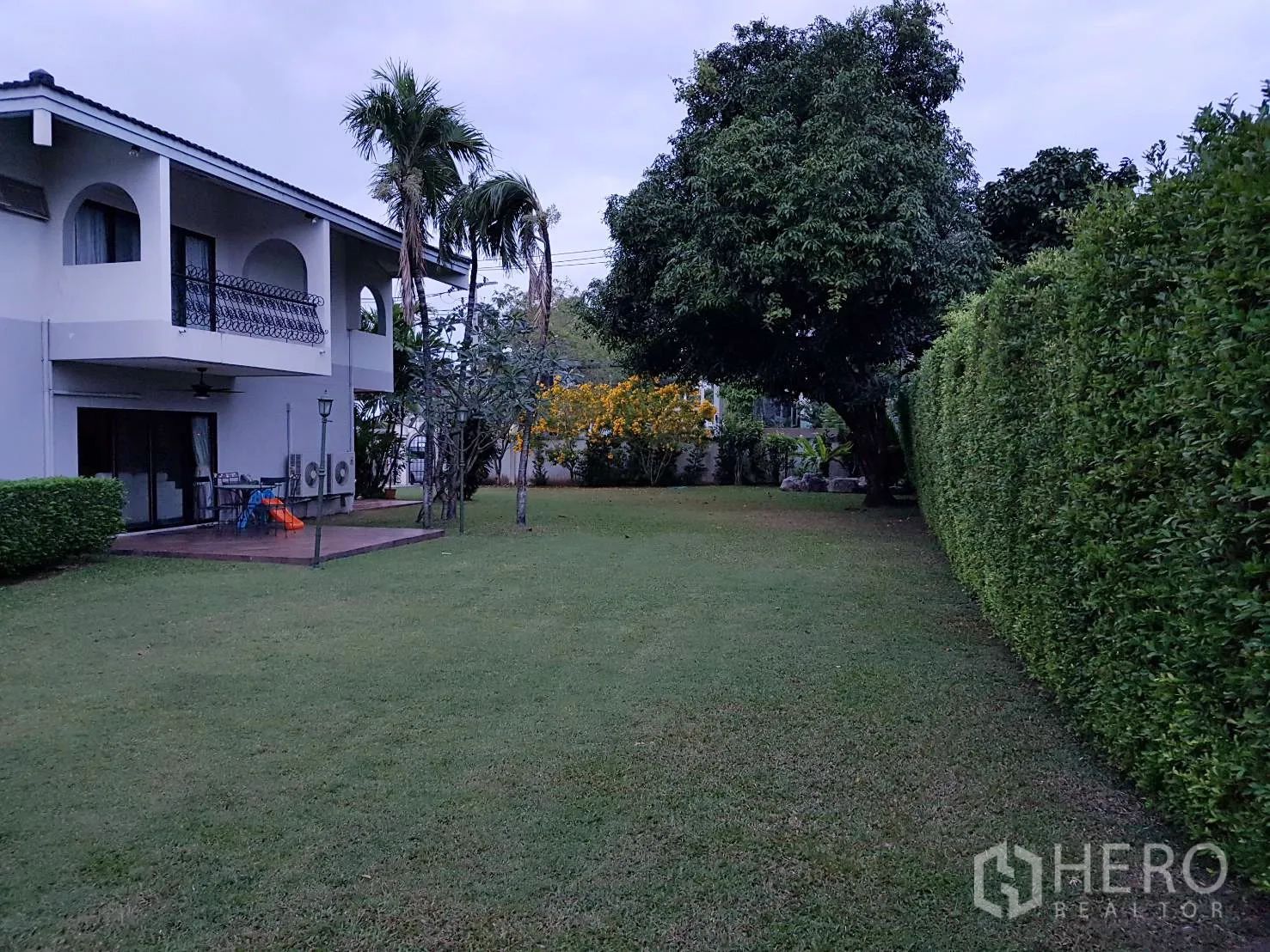 Detached House for rent in Khan Na Yao, Bangkok - Expansive private garden with manicured lawn, tall hedge and balcony view of a two-storey home.