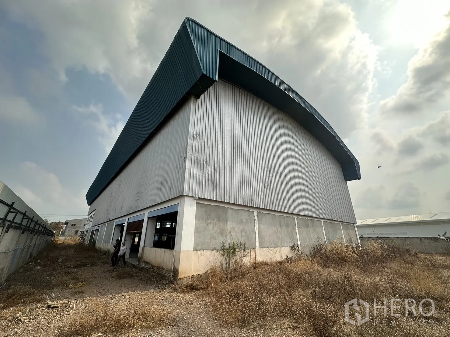 Factory for rent in Pak Kret, Nonthaburi - Side view of the warehouse’s corrugated metal wall and overhang with gravel service path.