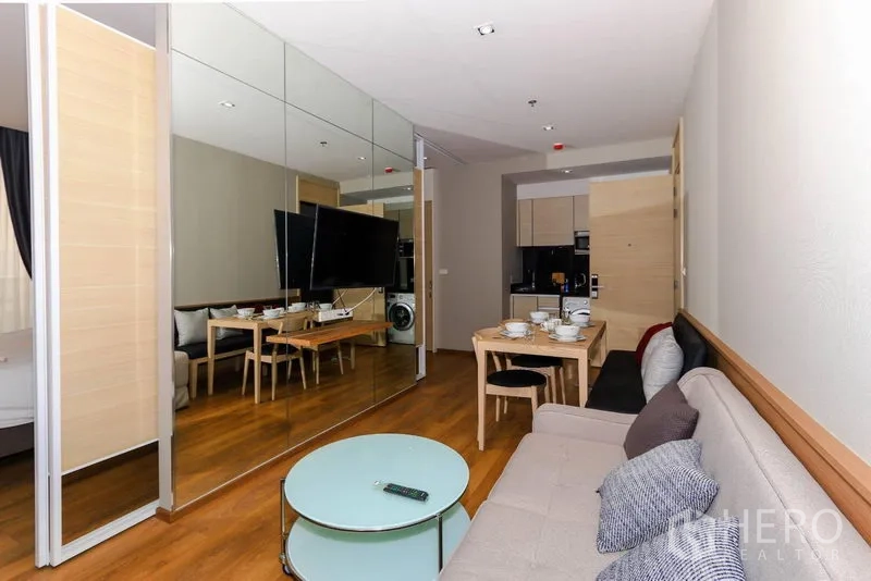 Condominium for rent in Khlong Toei, Bangkok - Open-plan living room with mirrored feature wall, mounted TV, dining set and warm wood flooring in a Bangkok condo.