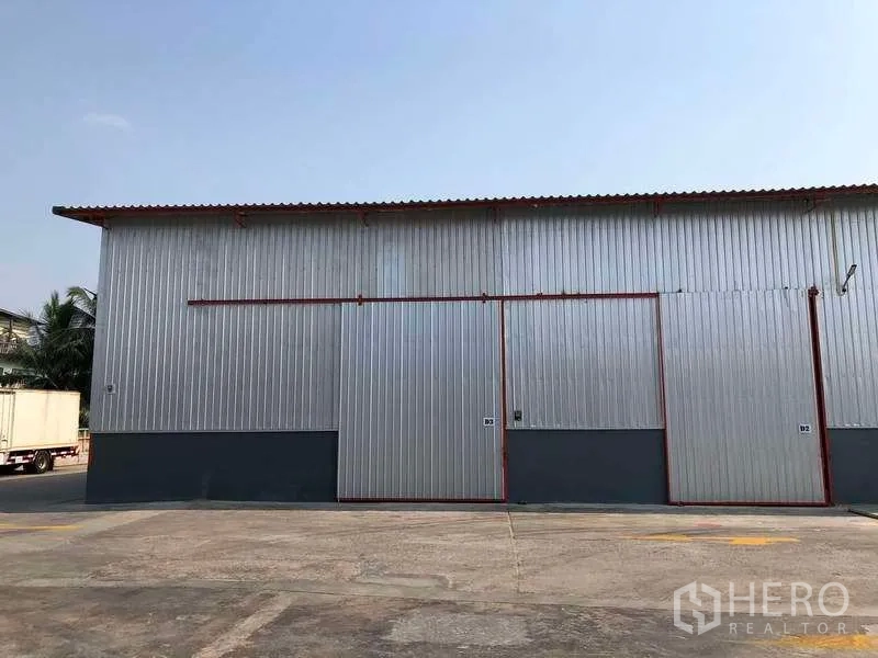 Factory for rent in Phra Pradaeng, Samut Prakan - Metal-clad factory exterior with large sliding doors and wide truck-friendly driveway under clear sky.