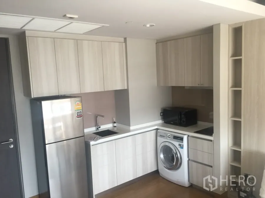Condominium for rent in Khlong Toei, Bangkok - Modern compact kitchen with light wood cabinets, fridge, microwave, cooktop, and washing machine.