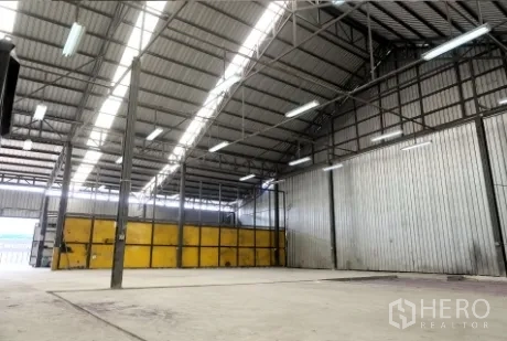 Warehouse for rent in Pak Kret, Nonthaburi - Open industrial space with corrugated metal walls, skylight strips, and large yellow sliding doors.
