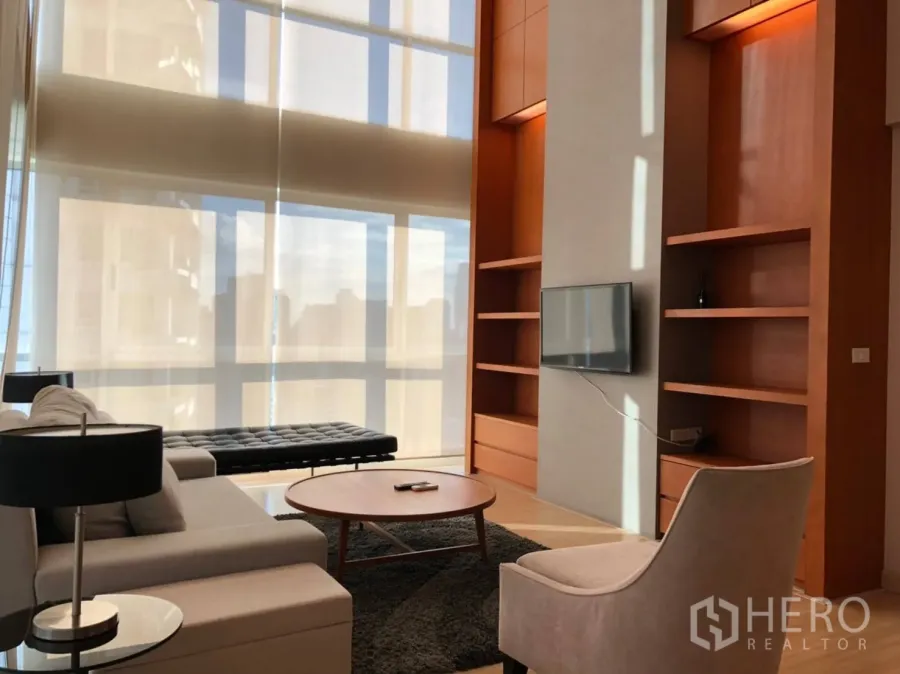 Condominium for rent in Pathum Wan, Bangkok - Bright living room with double-height windows, built-in shelves and a sofa set.