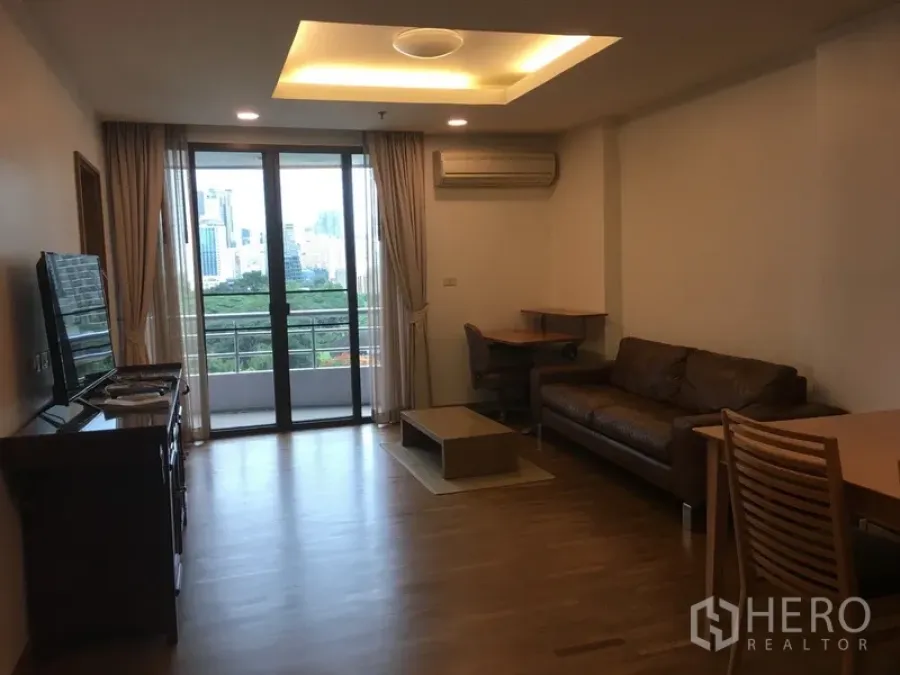 Condominium for rent in Pathum Wan, Bangkok - Living room facing open balcony with skyline and greenery view, brown sofa, TV stand and wood flooring.
