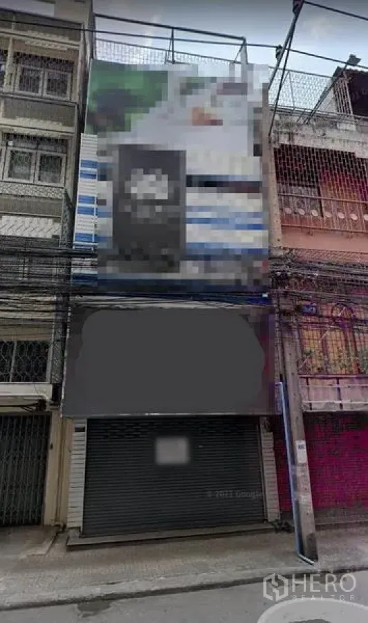 Shophouse for rent in Khlong Toei, Bangkok - Multi‑storey shophouse facade on a narrow Bangkok street with metal roller shutter, blank signage area, and overhead utility cables.