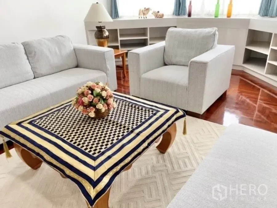 Condominium for rent in Khlong Toei, Bangkok - Bright living room with grey sofas around a wooden coffee table, built-in shelves and corner windows on hardwood flooring.