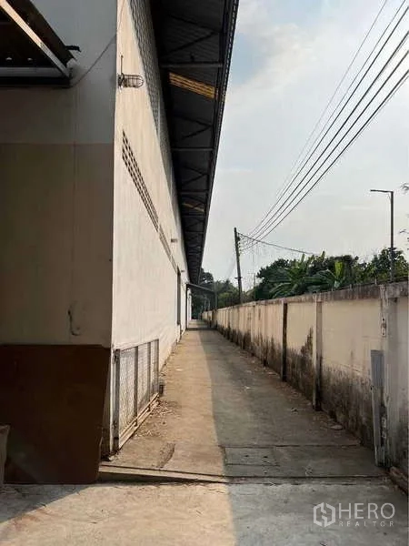 Warehouse for sale in Bang Bua Thong, Nonthaburi - Narrow concrete walkway running beside the warehouse with utility lines and fence.