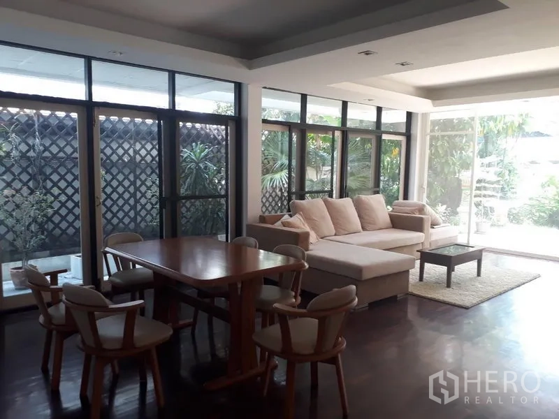 Detached House for rent in Watthana, Bangkok - Dining and lounge area with floor-to-ceiling glass opening to a green garden, sofa and wooden table
