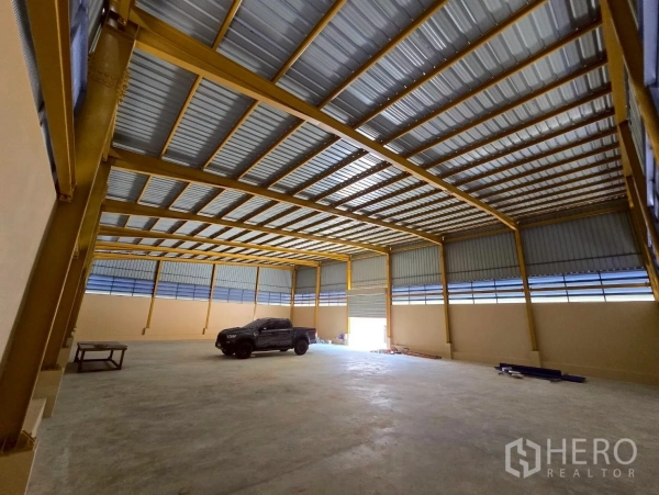 Warehouse for rent in Khlong Sam Wa, Bangkok - Spacious warehouse interior with high steel roof, yellow beams, concrete floor, and a wide roller shutter.