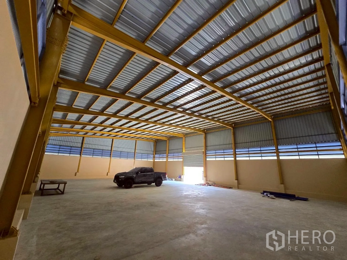 Warehouse for rent in Khlong Sam Wa, Bangkok - Spacious warehouse interior with high steel roof, yellow beams, concrete floor, and a wide roller shutter.