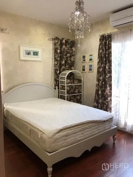 Detached House for rent or sale in Prawet, Bangkok - Guest bedroom with white bed, chandelier, floral curtains, and wood flooring.