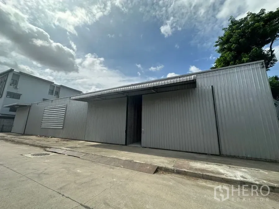 Warehouse for rent in Wang Thonglang, Bangkok - Street view of a single-storey metal warehouse with sliding steel doors and a covered loading awning.