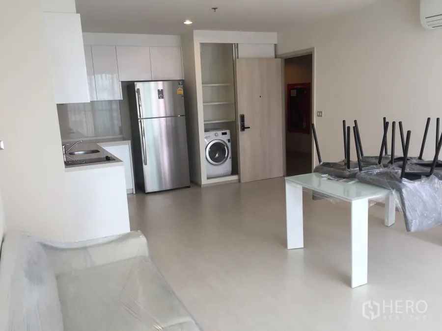 Condominium for rent in Khlong Toei, Bangkok - Open-plan living area with white kitchen, stainless refrigerator, and in-unit washer in a modern Bangkok condo.