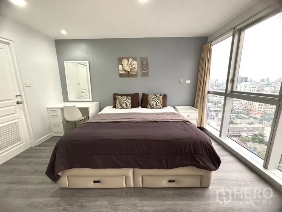 Condominium for sale in Khlong Toei, Bangkok - Bright master bedroom with king bed, grey accent wall, desk, and floor‑to‑ceiling city‑view windows.