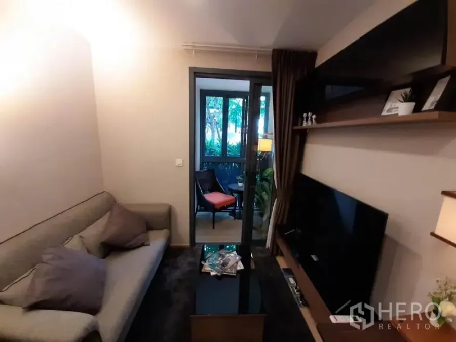 Condominium for rent or sale in Watthana, Bangkok - Cozy living room with sofa, TV wall and sliding glass door opening to a balcony with greenery.