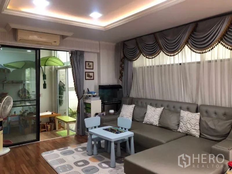 Detached House for sale in Lam Luk Ka, Pathum Thani - Family lounge with large sectional sofa, kids play area and sliding glass doors to a sunroom.