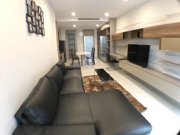 Condominium for rent in Pathum Wan, Bangkok - Open-plan living room with black leather sofa, wall-mounted TV and built-in shelving.