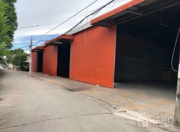 Warehouse for rent in Bang Kapi, Bangkok - Street view of a red metal-clad warehouse with multiple loading bays and a wide driveway in Bang Kapi.