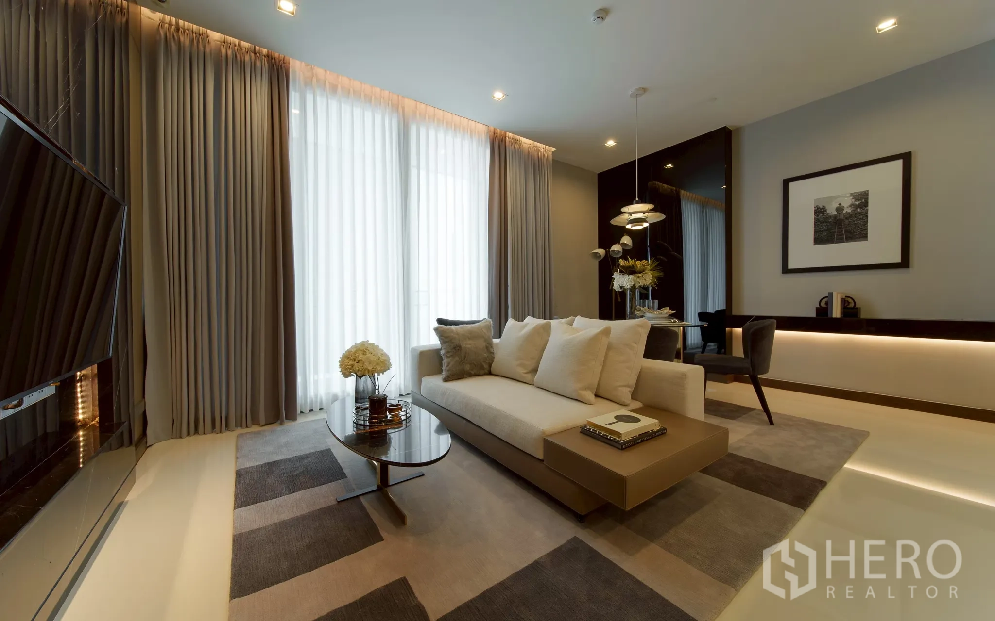 Condominium for sale in Khlong Toei, Bangkok - Modern living and dining area with neutral sofa, coffee table, floor-to-ceiling curtains, pendant lights, wall-mounted TV, and warm ambient lighting.