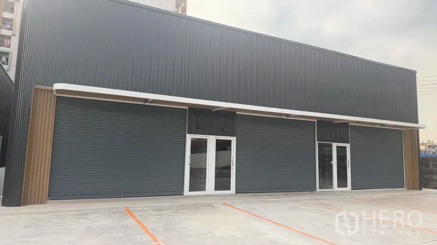 Warehouse for rent in Prawet, Bangkok - Modern warehouse exterior with dark metal cladding, glass doors, roller shutters and marked parking bays.