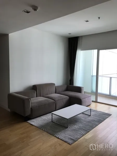 Condominium for rent in Khlong Toei, Bangkok - Bright Sukhumvit condo living room with gray L-shaped sofa, white coffee table, rug and balcony access.