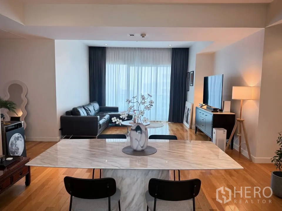 Condominium for rent or sale in Khlong Toei, Bangkok - Open-plan dining with a marble table leading to a living room with a leather sofa, TV unit, and sheer curtains.