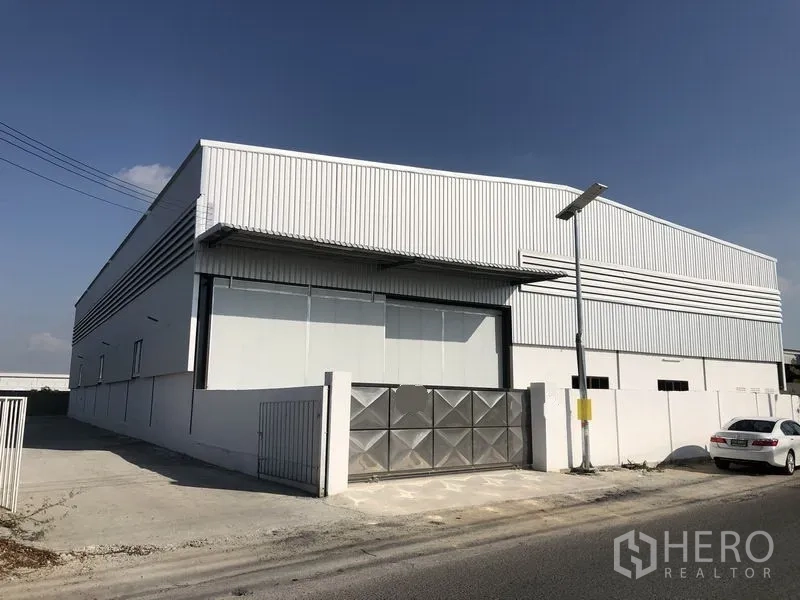 Factory for rent in Bang Phli, Samut Prakan - Modern metal-clad factory exterior in Bang Phli with high roof, large roller shutter, gated entrance and concrete driveway on a wide road.