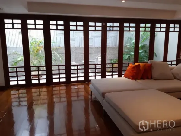 Townhouse for rent in Watthana, Bangkok - Sunlit living room with floor-to-ceiling wooden-framed glass doors opening to a private patio with potted palms and a white sectional sofa.