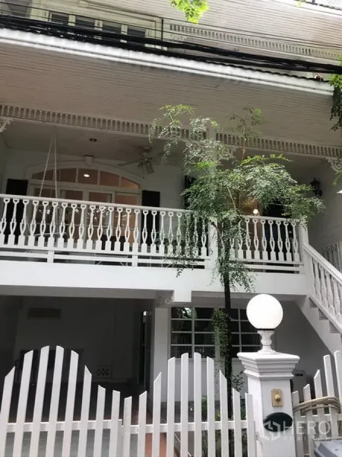 Townhouse for rent in Mueang Samut Prakan, Samut Prakan - White two-storey townhouse exterior with balcony, decorative railings, gated entry and leafy tree.