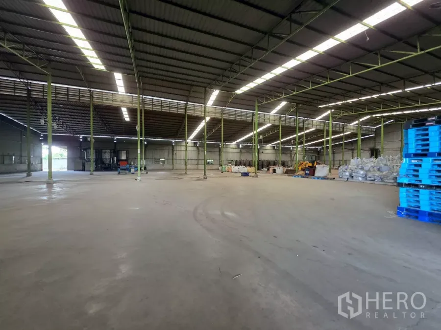 Factory for rent in Mueang Samut Sakhon, Samut Sakhon - Spacious warehouse interior with high steel truss roof, skylight strips and wide concrete floor with storage and pallets.