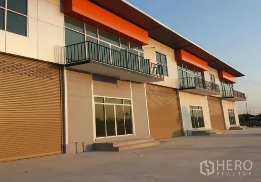 Factory for rent in Pathum Thani - Modern factory frontage with balconies, glass entry, and multiple roll-up doors at sunset.